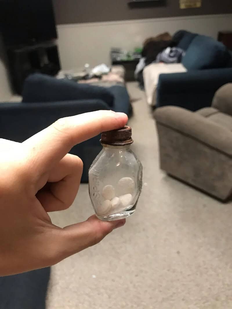 Hand holding a small glass jar with a brown lid, containing white pills, in a living room.