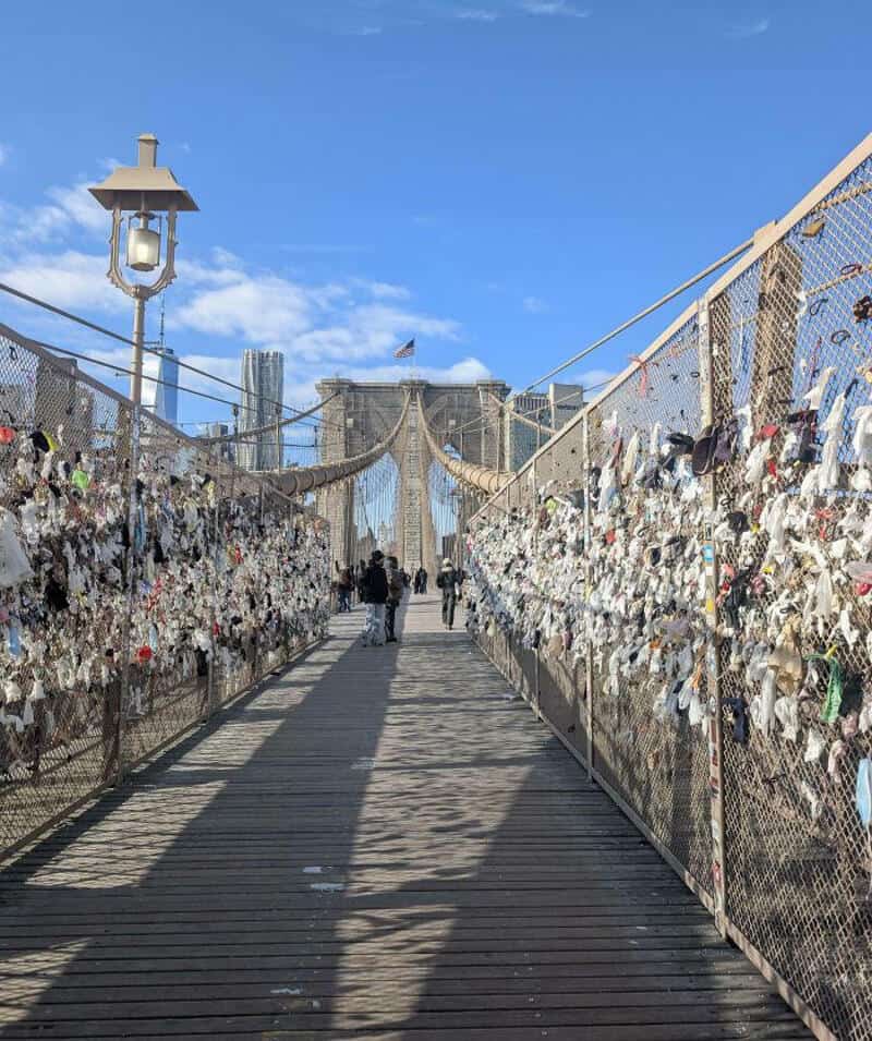 Bridge with padlocks and people walking, Brooklyn Bridge in background, clear sky.