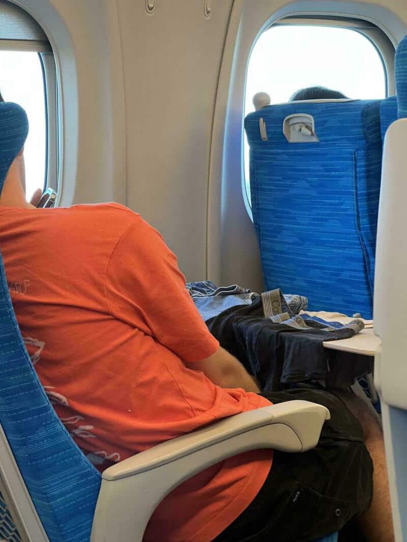 Passenger in orange shirt sitting on airplane with blue seats and window view.