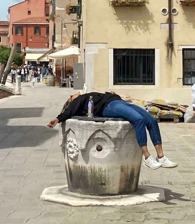 Person lying across a large stone fountain in a city square.