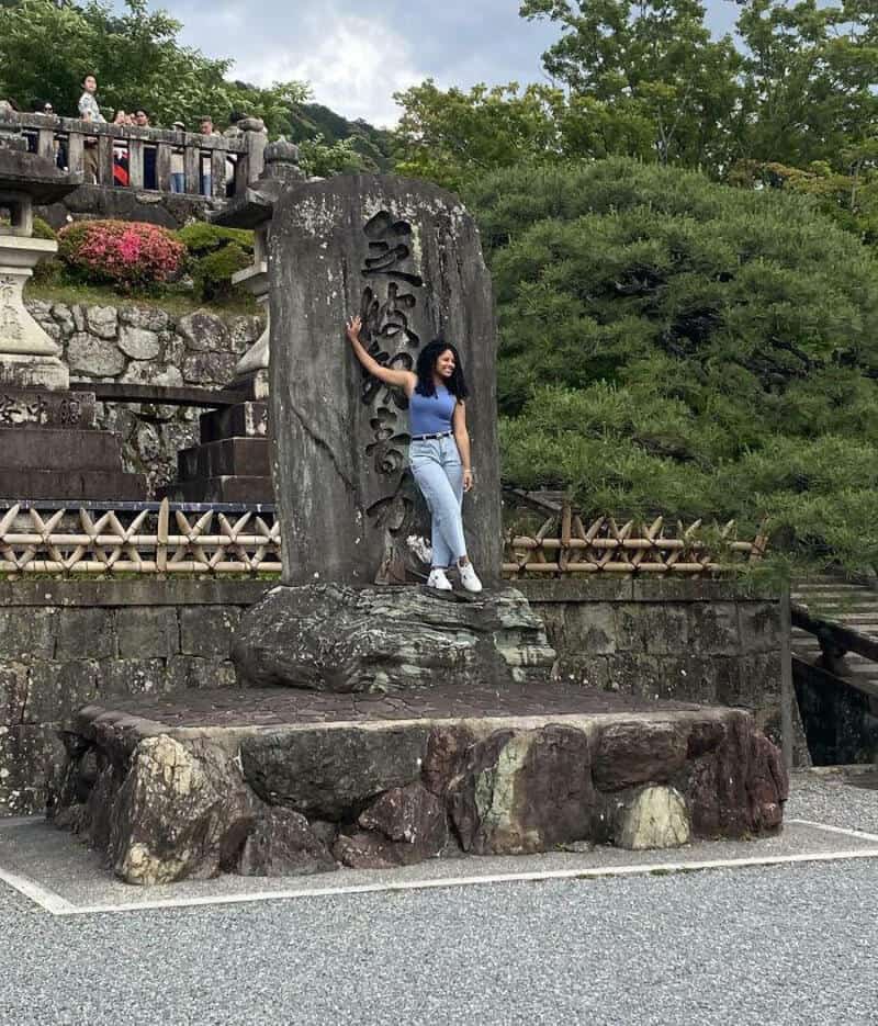 Woman in blue top and jeans leaning on a large stone monument in a park.