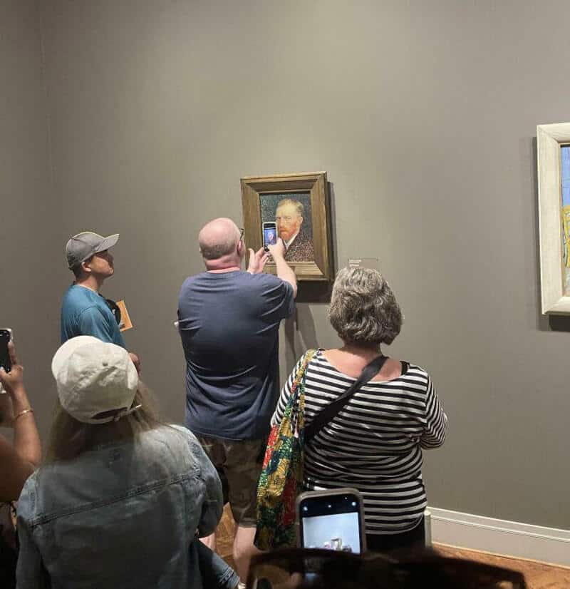Tourists taking photos of a painting in a museum, some with phones and cameras.