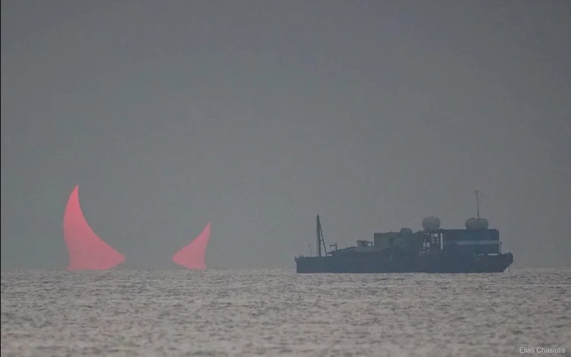 Two pink sailboats in the distance with a ship nearby on the water.