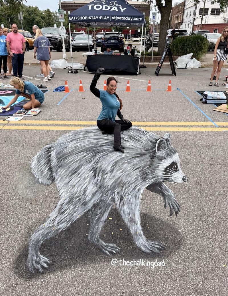 Woman sitting on a giant raccoon street art with a black box on her head.