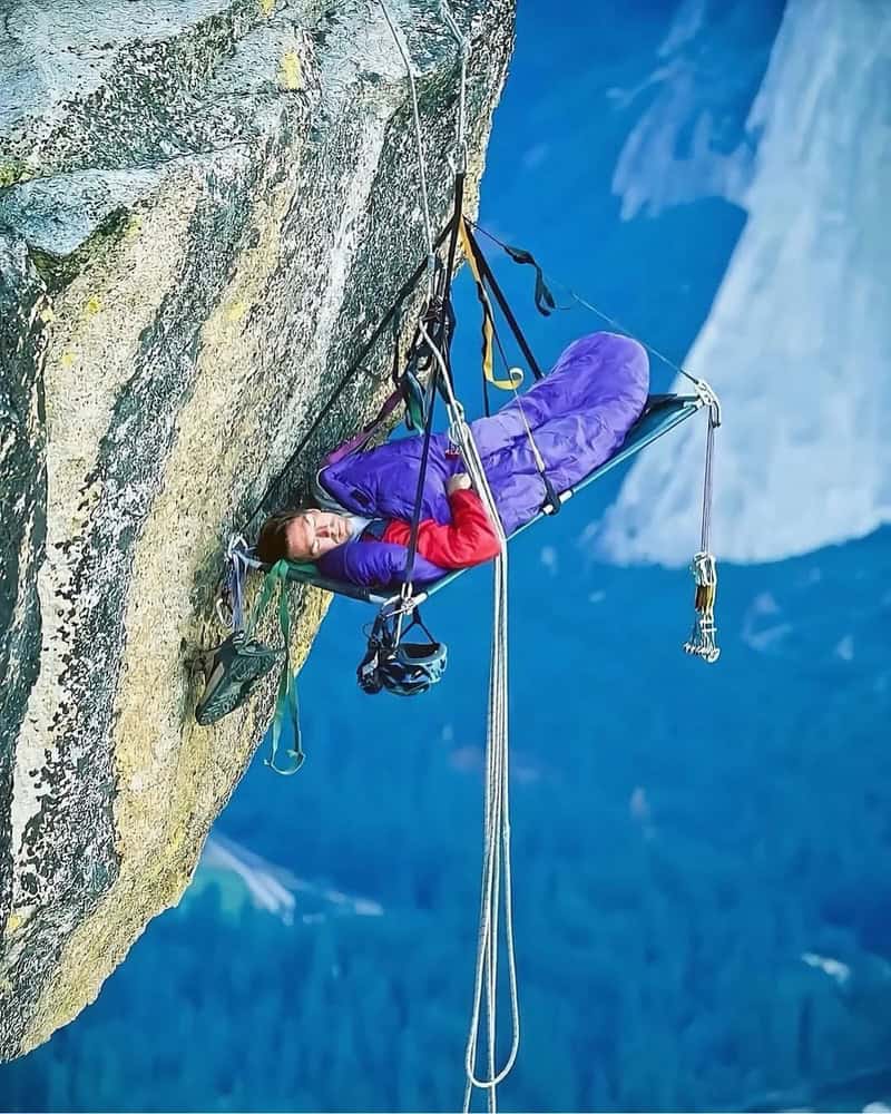 Climber resting on a ledge during a mountain climb with a harness and ropes.