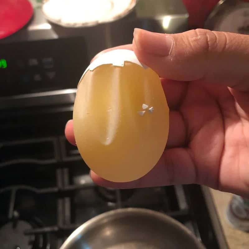 Hand holding a cracked eggshell over a stove with a pot below.