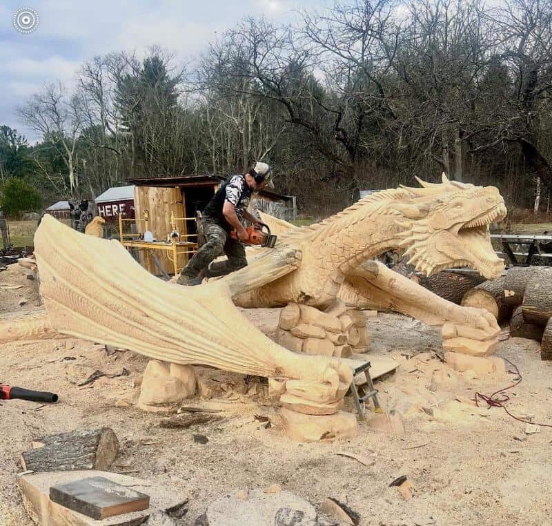 Carver working on a large wooden dragon sculpture outdoors.