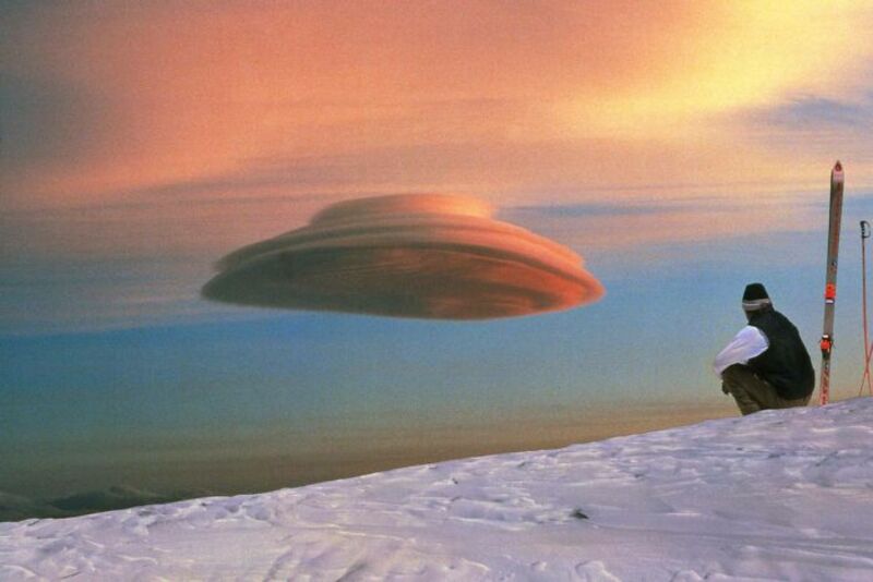 Person sitting on snow with UFO-shaped cloud in sky during sunset.