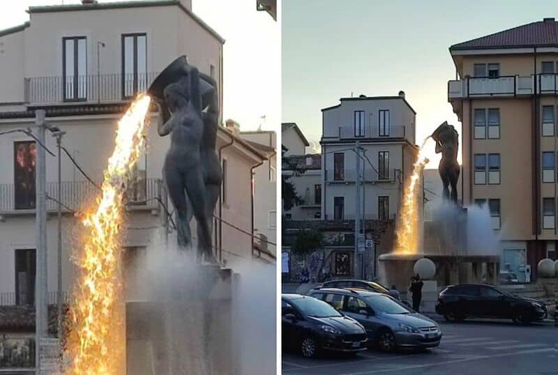 Statue with fire and water effects in front of buildings, city scene.