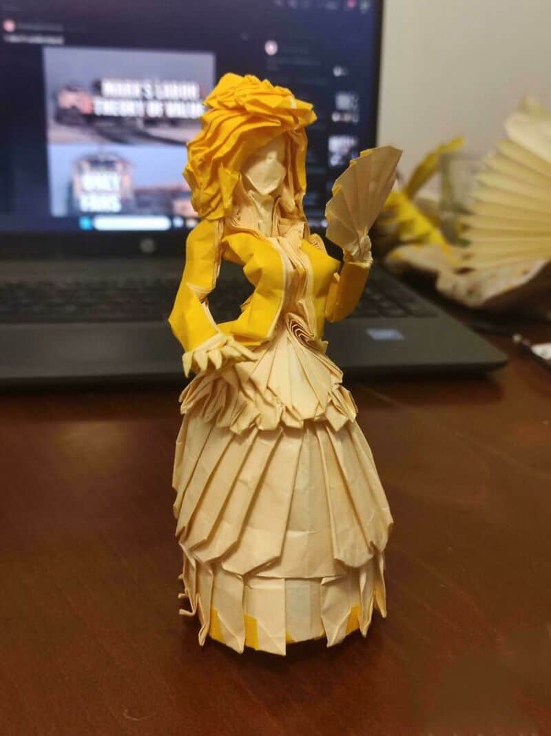 Origami woman with yellow hair and dress holding a fan, on a desk with a laptop.