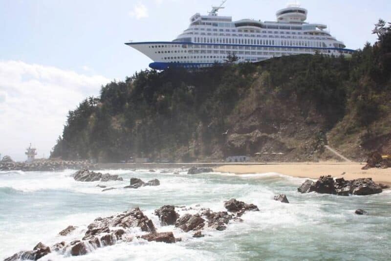 Large cruise ship near rocky beach with trees and cliffs.