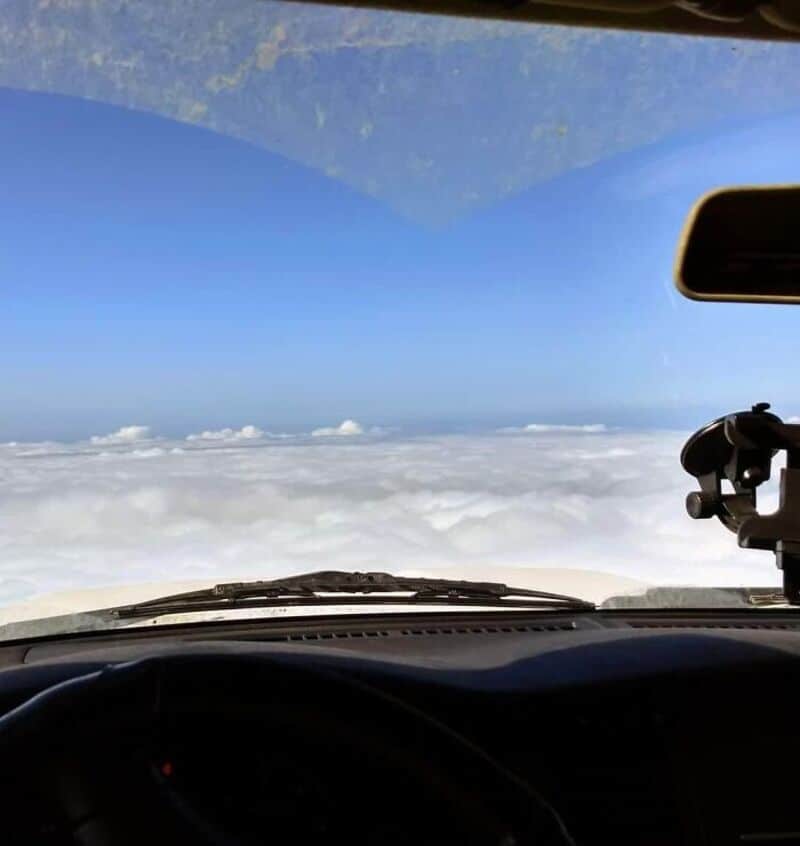 View from airplane cockpit above clouds with blue sky and white clouds.