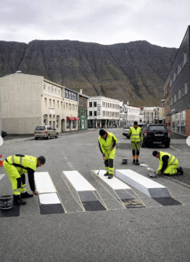 Four workers in yellow vests painting a crosswalk on a street with buildings and mountains in the背景.