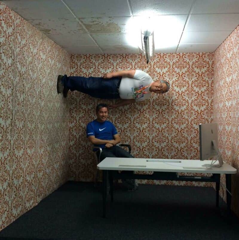 Two men in an office with patterned wallpaper, one standing on the ceiling and one sitting at a desk.