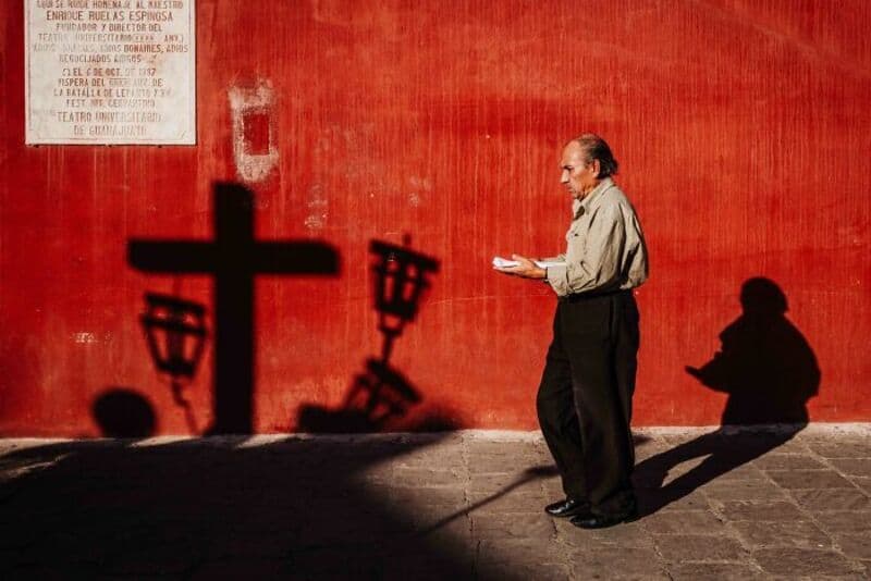 Older man reading a paper against a red wall with shadows of street lamps.