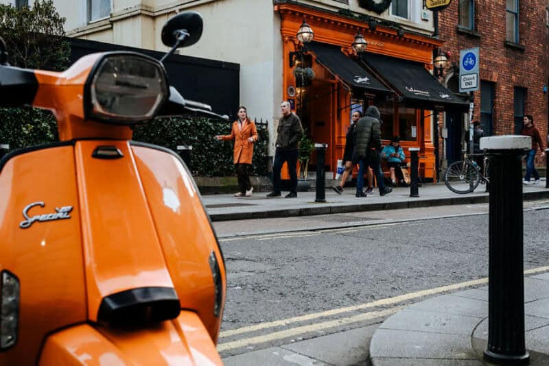 Orange scooter parked on the street with people outside a cafe.