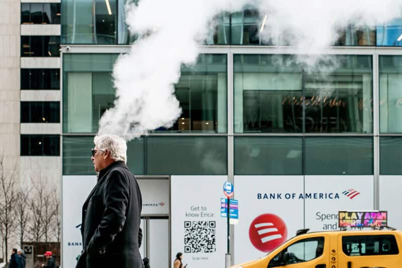 Older man walking past a building with steam rising from a manhole.