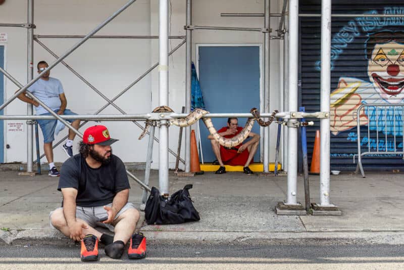 Man sitting on sidewalk with clown mural behind, others nearby, scaffolding present.