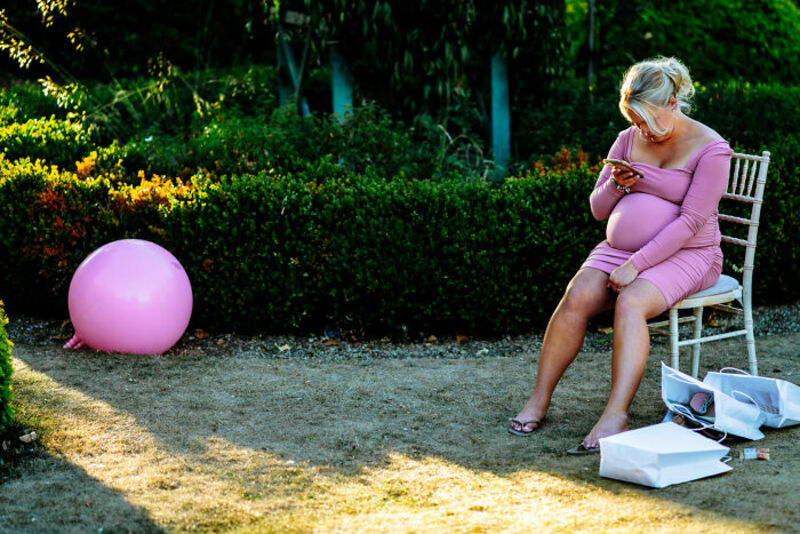 Pregnant woman in pink dress sitting on a chair and looking at her phone in a garden.
