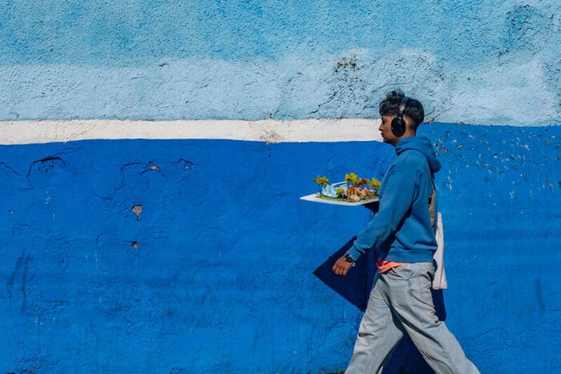 Young man with headphones walking past a blue wall with a white stripe.