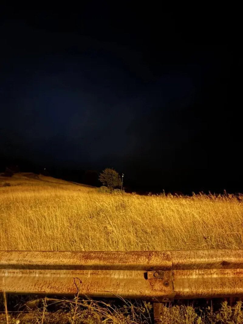 Dark night sky over a grassy field with a tree and a guardrail in the foreground.