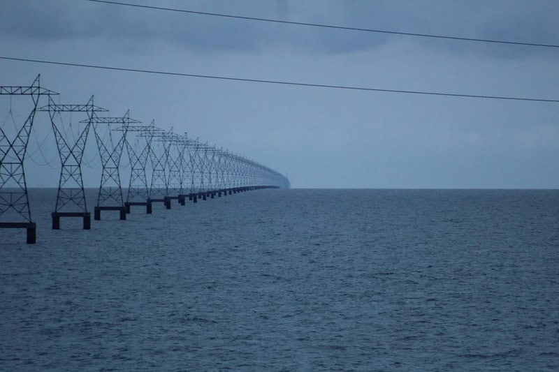 Power lines over water under a cloudy sky, stretching into the distance.
