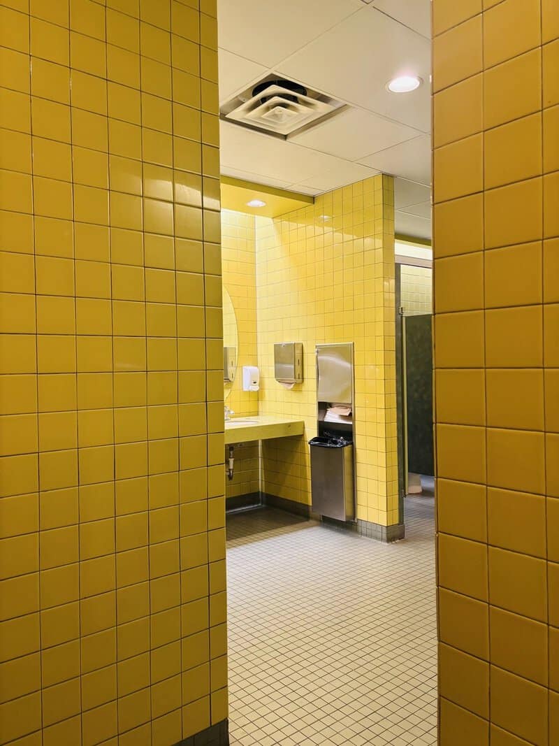 Empty yellow-tiled bathroom with a sink, paper towel dispenser, and trash bin.
