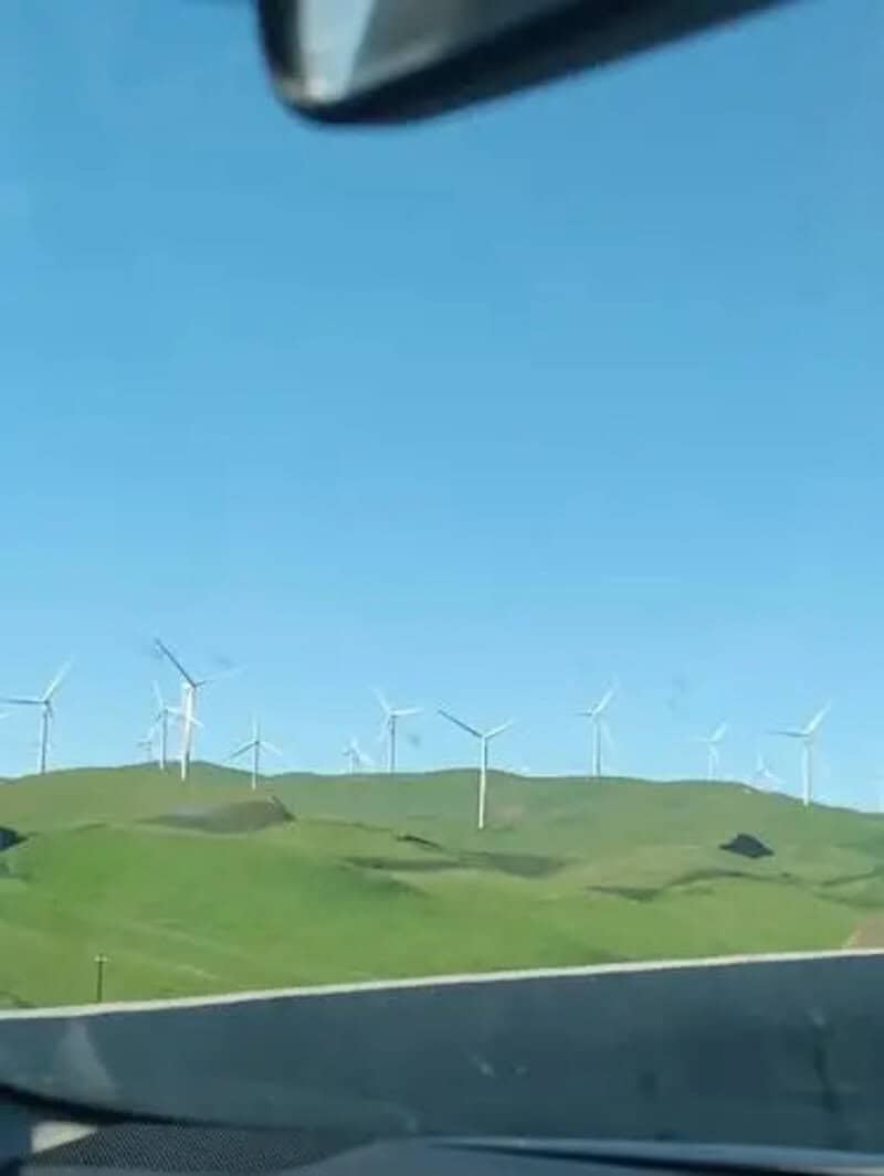 Wind turbines on green hills under a blue sky, seen from a car window.