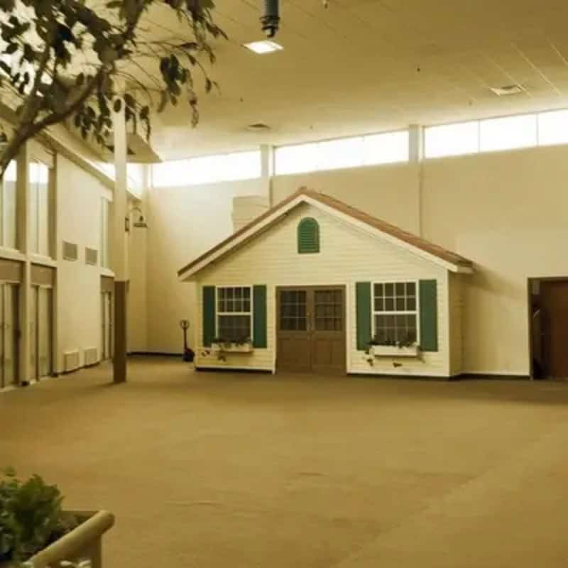 Small house model inside a large, empty room with high ceiling and windows.