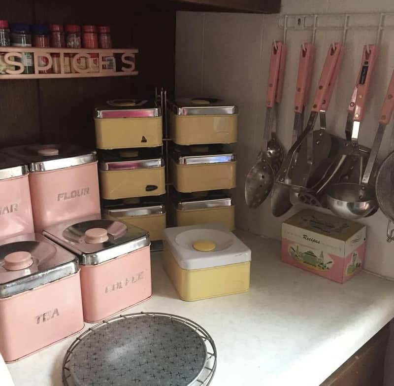 Vintage kitchen storage with pink and yellow tins, utensils, and a wire rack.