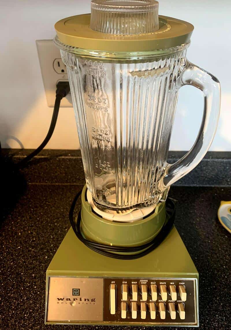 Vintage blender with glass pitcher and green base plugged into wall socket.