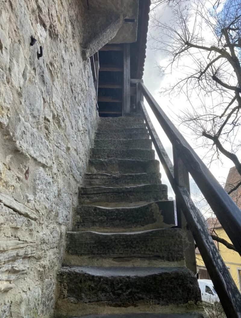 Old stone staircase with wooden railing leading up to a door under a roof.