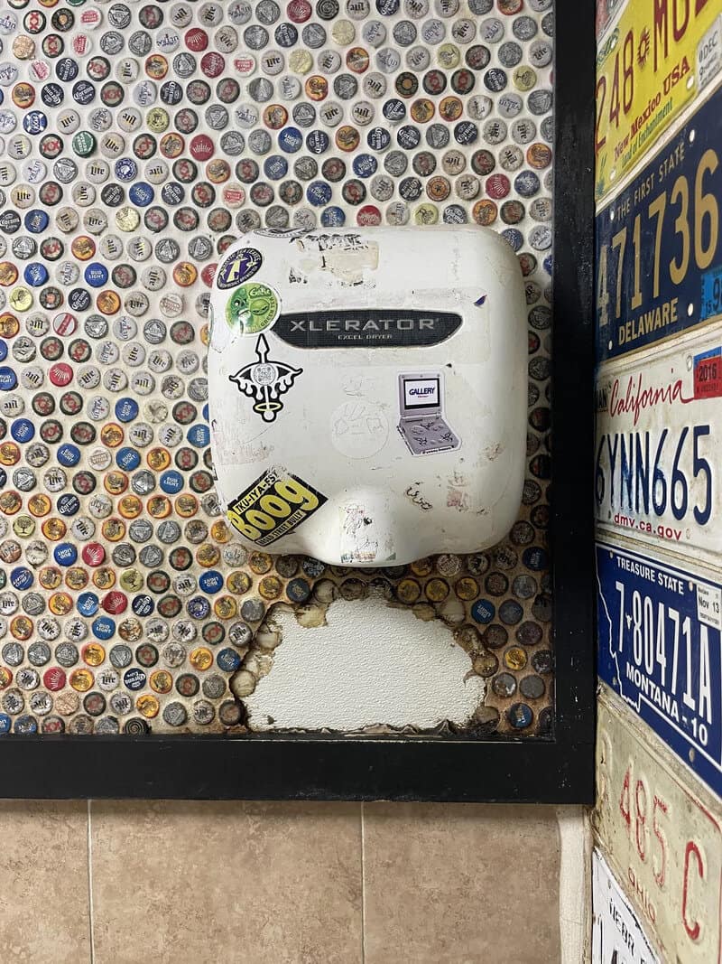 Old Xlerator hand dryer with stickers on a wall of bottle caps and a sign with phone numbers.