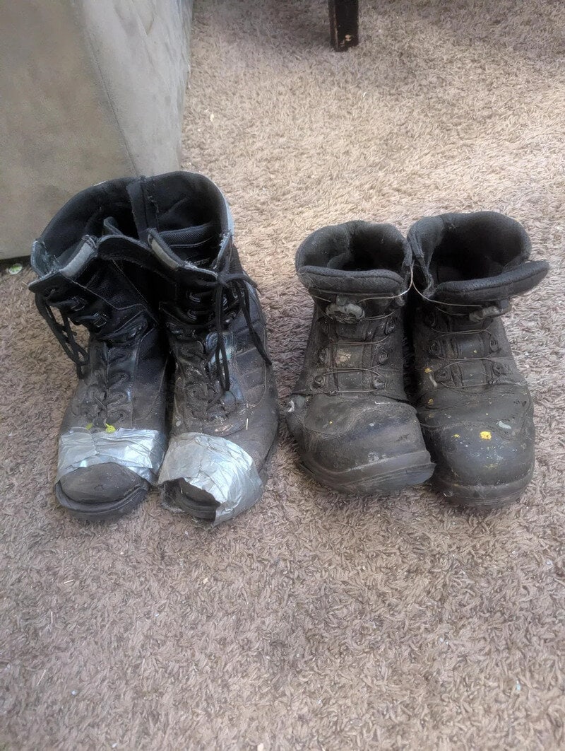 Pair of worn, dirty black work boots on a carpeted floor.