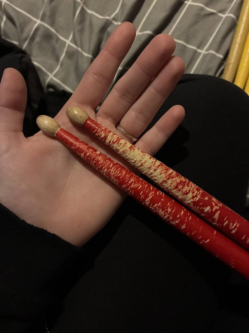 Person holding two red and beige drumsticks with a hand and a bed in the background.