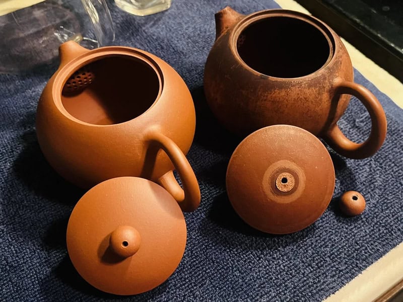 Two unglazed clay teapots with lids and a small clay ball on a dark fabric surface.