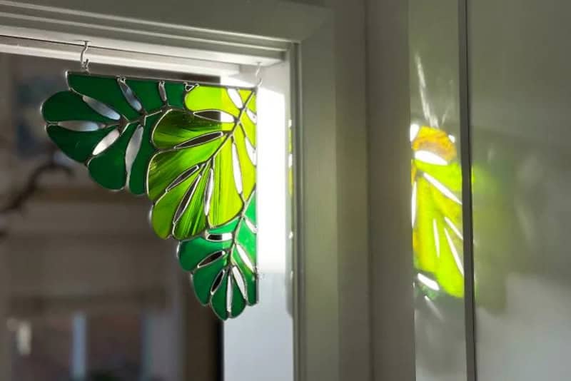 Green monstera leaf-shaped stained glass hanging on window with sunlight.
