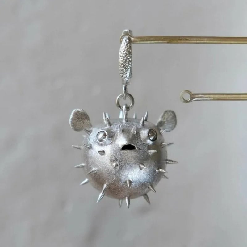Silver pufferfish pendant with spikes, eyes, and open mouth, hanging from a pin.