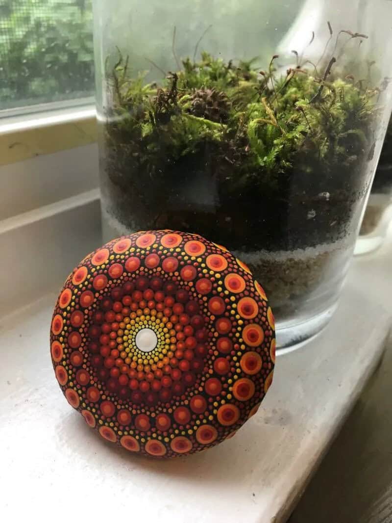 Colorful mandala painted rock with moss in a glass container on a windowsill.