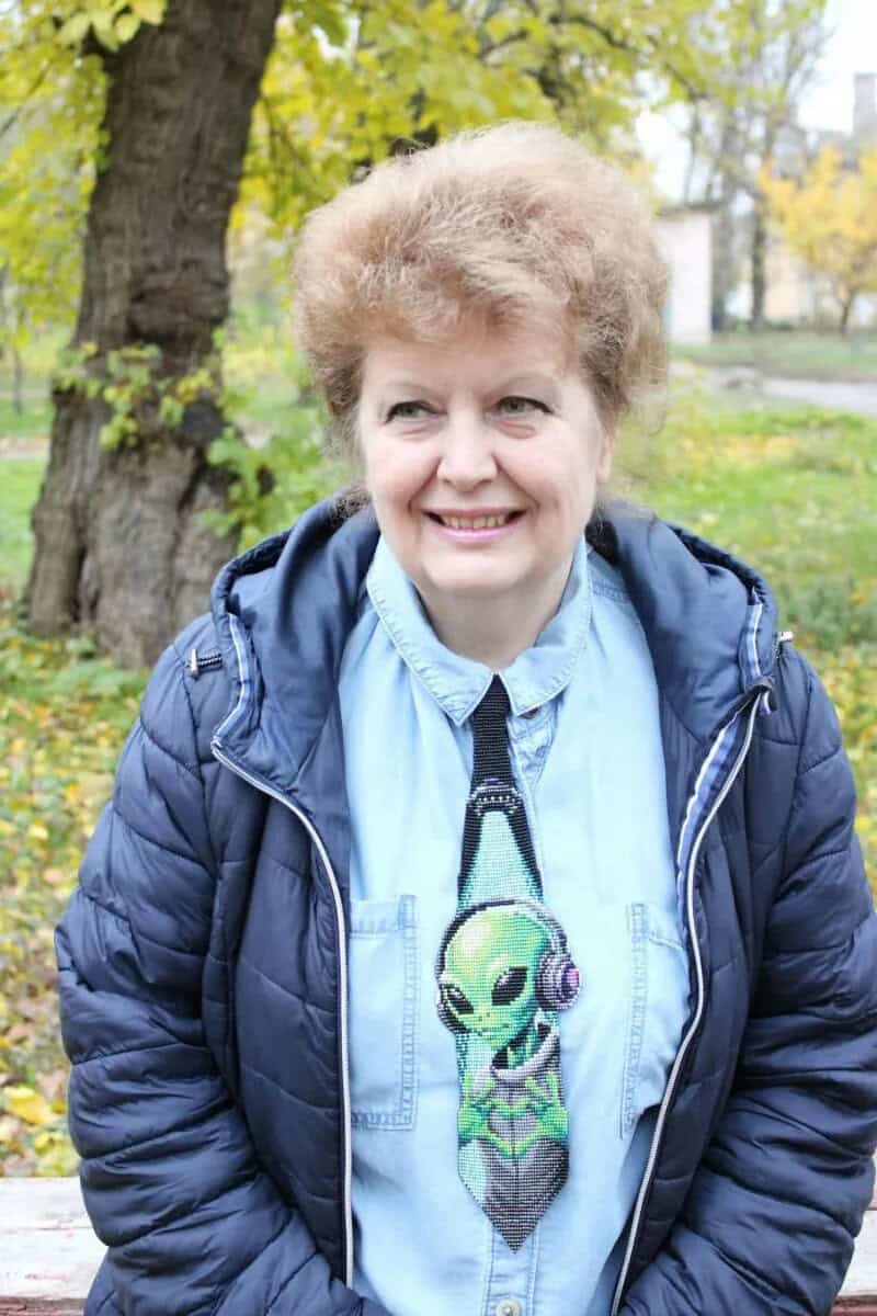 Smiling woman outdoors wearing a blue jacket and a necklace with an alien pendant.