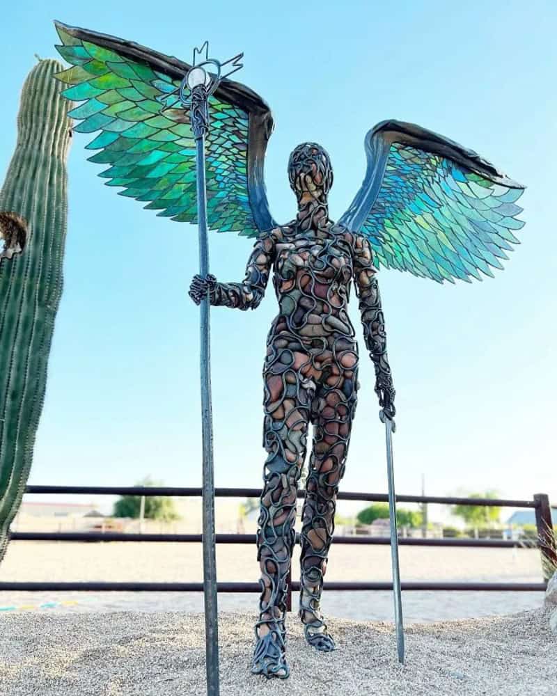 Metal sculpture of a winged figure with colorful wings and a staff, outdoors.