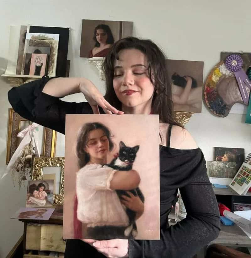 Young woman holding a portrait of herself with a black and white cat in an art studio.