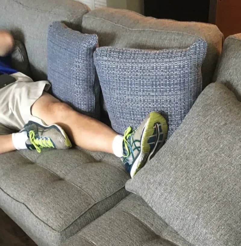 Child's legs and feet on a gray couch with cushions, wearing sneakers and shorts.