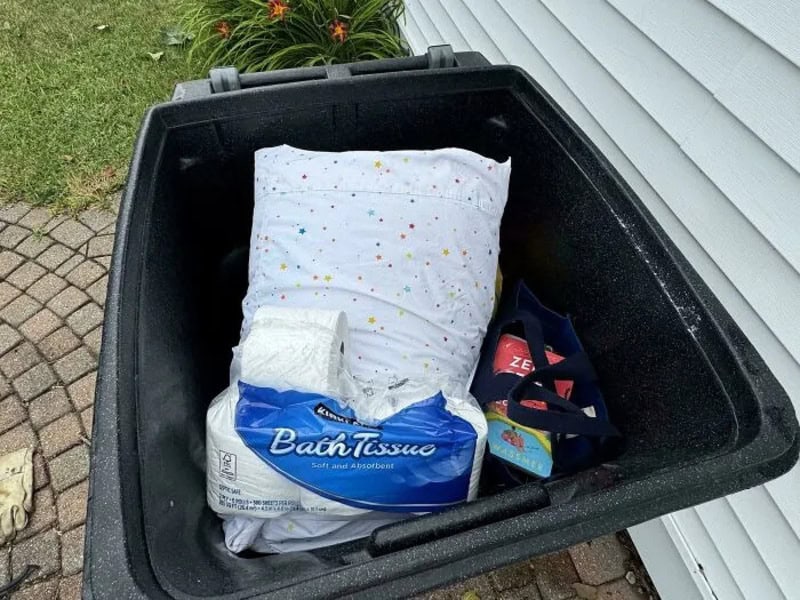 Trash bin with a pillow, toilet tissue, and cleaning supplies inside.