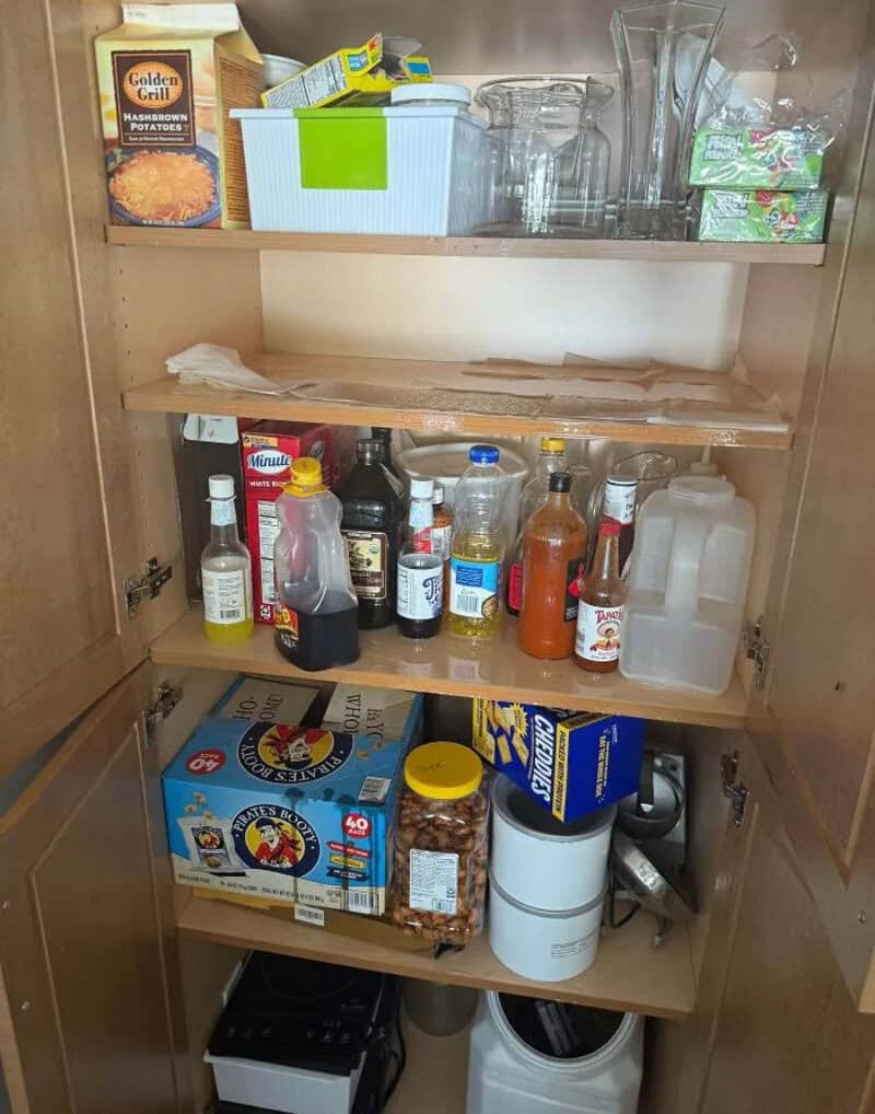 Open kitchen cabinet with condiments, snacks, and glasses inside.