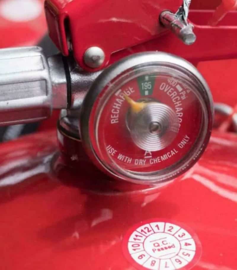 Fire extinguisher pressure gauge showing overcharge, attached to a red fire safety device.