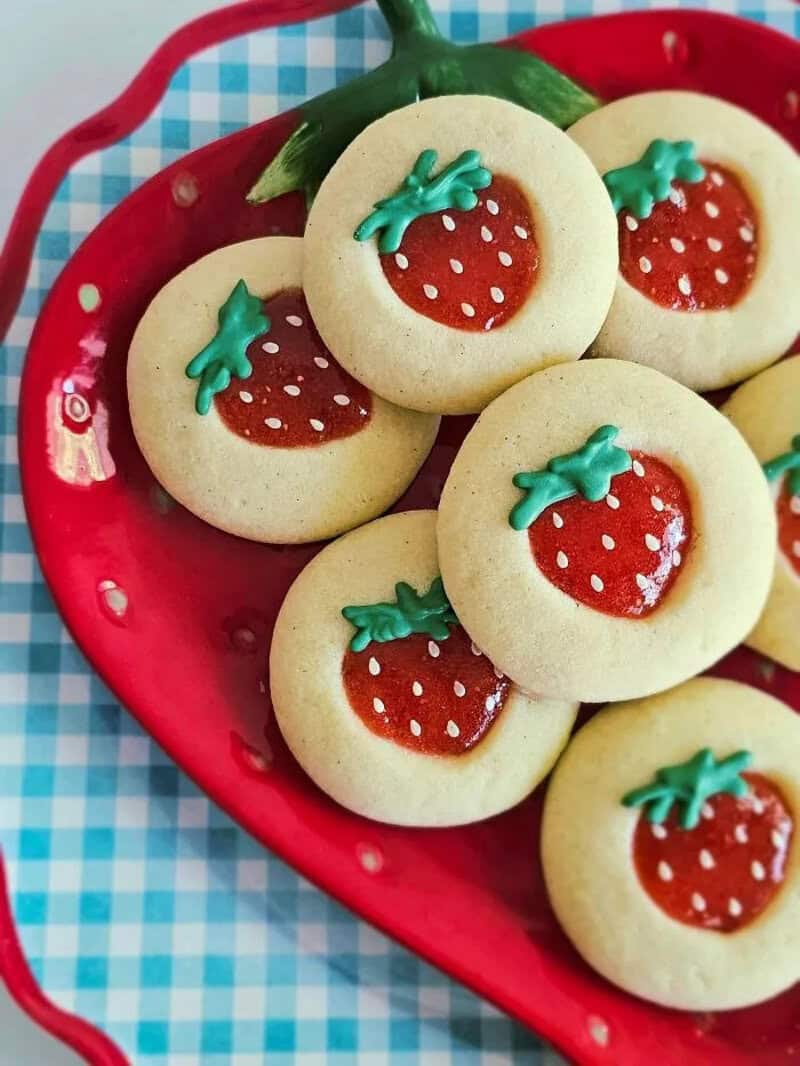 Cookies decorated as strawberries in a red bowl on a blue checkered cloth.