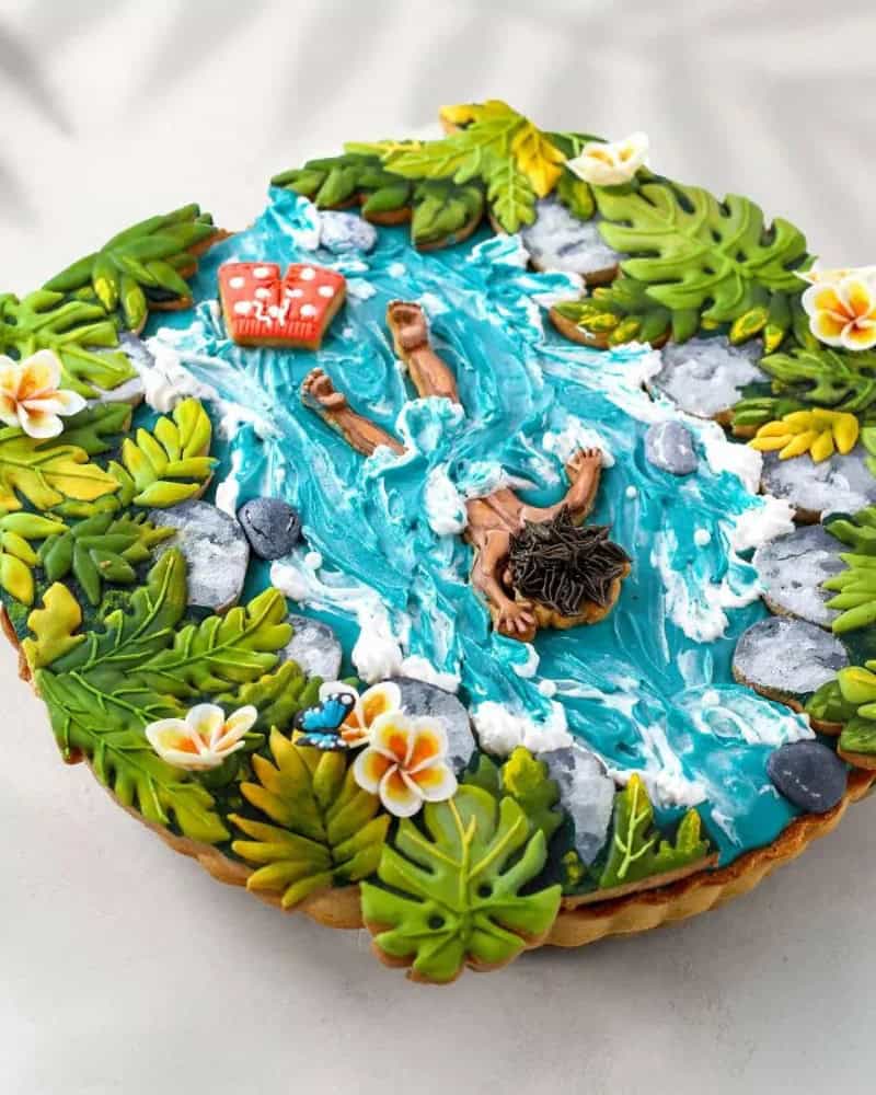 Cake decorated as a swimming pool with a swimmer and tropical plants.