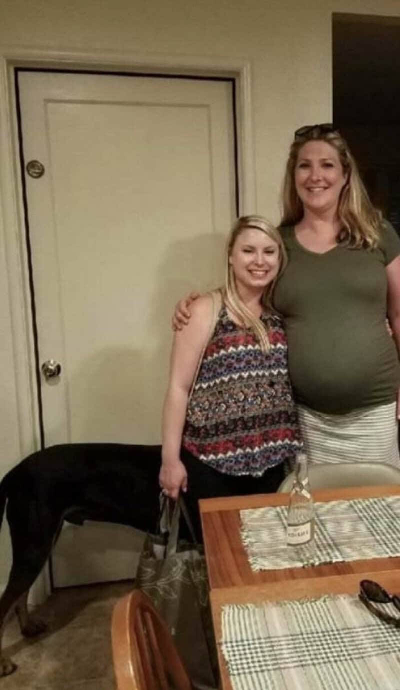 Two women, one pregnant, standing together in a room, smiling at the camera.
