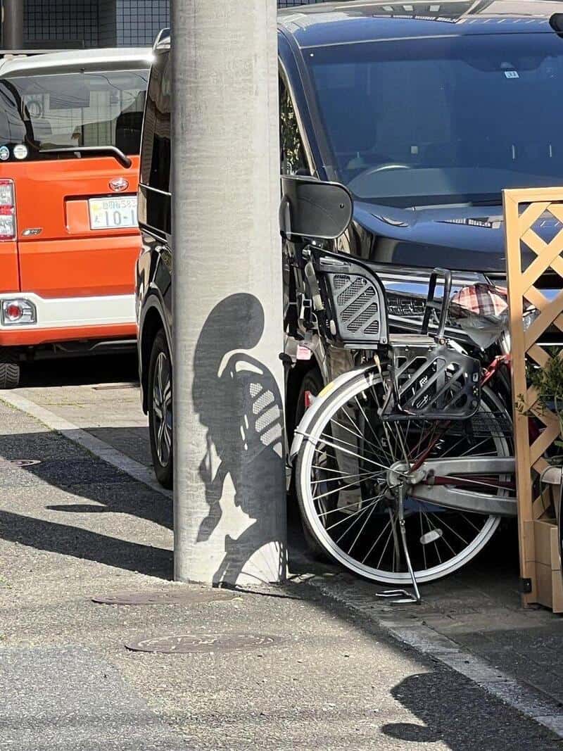 Shadow of a person riding a bicycle cast on a pole, with cars and a fence nearby.